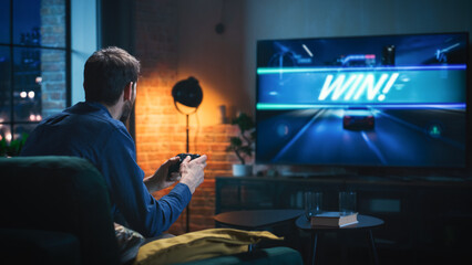 Young Man Spending Time at Home, Sitting on a Couch in Stylish Loft Apartment and Playing Arcade Car Video Games on Console. Male Using Controller to Play Street Racing Drift Simulator. © Gorodenkoff