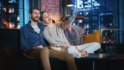 Portrait of Beautiful Couple Spending Time at Home, Sitting on a Couch, Hugging and Watching Exciting TV Show in Their Stylish Loft Apartment. Man and Woman Streaming Comedy Movie and Have Good Time.