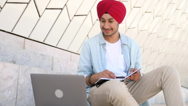 Indian student man in traditional headwrap sitting outdoors and using laptop for learning on the distance, hindu guy watching webinar and taking notes, male student studying online, e-learning