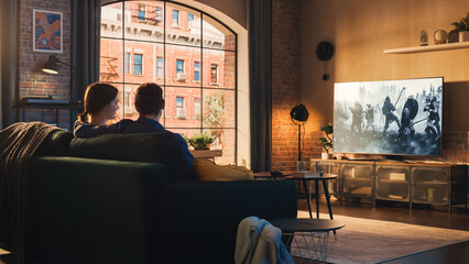 Authentic Couple Spending Time at Home, Sitting on a Couch and Watching Latest Blockbuster on Flat...