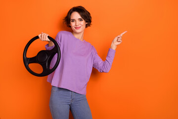 Portrait of pretty positive girl hold wheel direct finger empty space offer isolated on orange...