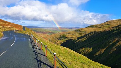 View of the empty road and a rainbow above green hills. Buttertubs Pass, Yorkshire Dales, England.