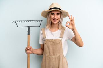 blonde woman feeling happy, showing approval with okay gesture. farmer and rake cocnept