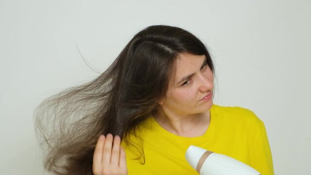 A brunette woman dries her hair with a hair dryer, looks at her hair with a dissatisfied look