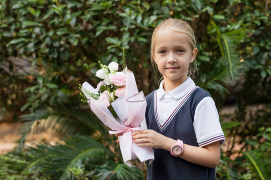 Lifestyle portrait of Little caucasian girl with blonde hair six-seven years old from elementary school outside. Happy Kid with bouquet of flowers is going learn new things 1th of september after end