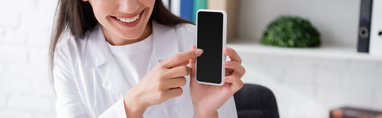 Cropped view of smiling doctor pointing at smartphone with blank screen in clinic, banner