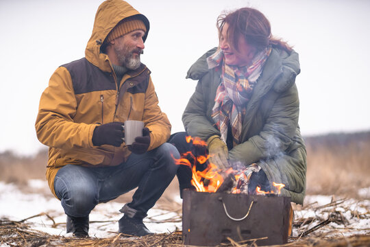 Warming up at campfire middle aged man and woman away in the wild drinking hot tea or coffee. Mature couple making a barbecue in winter forest at camp fire. Family journey. Family vacation concept. 