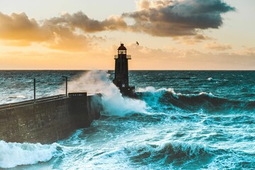 Large waves crash against the stone tower of the lighthouse at high tide at sunset - obrazy, fototapety, plakaty