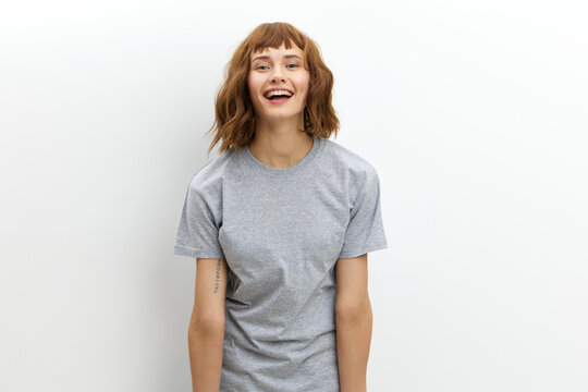 a happy, joyful woman in a gray cotton T-shirt stands against a light background, smiling happily and looking at the camera. Horizontal photo with an empty space for inserting an advertising layout
