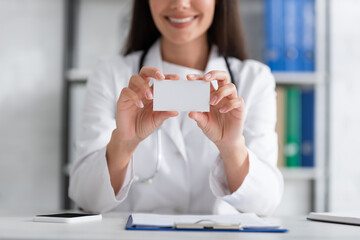 Cropped view of smiling blurred doctor holding empty business card near smartphone and clipboard in...