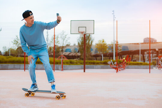 Young hipster guy skateboarder selfie with mobile phone while sitting on skateboard outdoors