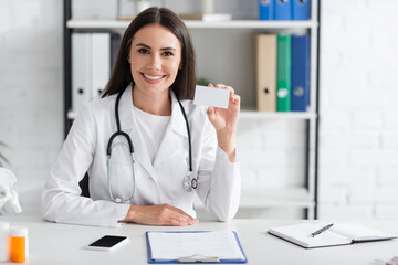 Smiling doctor holding business card with copy space near smartphone and clipboard in clinic