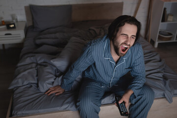 Tired man yawning and holding remote controller in bedroom at night