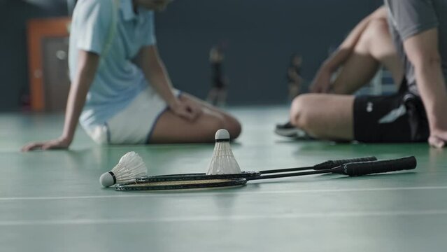 Ground level close up shot of two rackets and shuttlecocks on floor of indoor badminton court. Unrecognizable male and female athletes chatting in defocused background