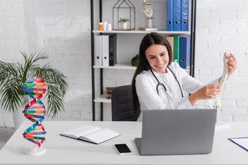 Positive doctor holding spinal model during online consultation on laptop near notebook and...