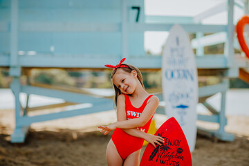 Pretty little girl in red bikini posing with small surfboard like a model on the beach against blue lifeguard tower