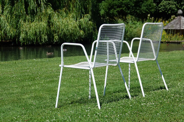 Zwei weisse Metallst&uuml;hle auf einer gr&uuml;nen Wiese vor einem Ententeich