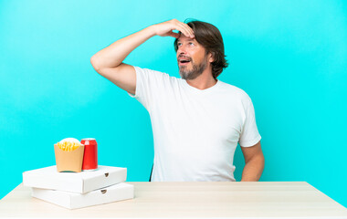 Senior dutch man eating fast food sitting in a table doing surprise gesture while looking to the side