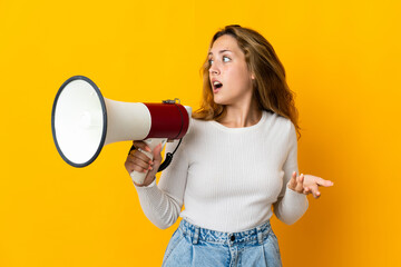 Young blonde woman isolated on yellow background holding a megaphone and with surprise facial...