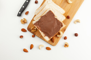 Square bread for toast with chocolate paste on a wooden board. Nuts, a knife and a wooden cutting board with a sandwich and slices of bread on a white table.