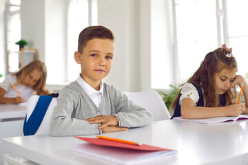 Boy get ready to start study. Portrait of cute smiling little schoolboy sitting in classroom with his arms folded flat on desk. Male elementary school student is looking at camera. Education concept.
