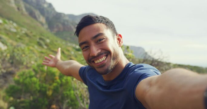 Portrait of a happy man hiking and pointing to the scenic views while taking selfies and making a video call. Face a young Latino tourist smiling and laughing while exploring during a trek in nature