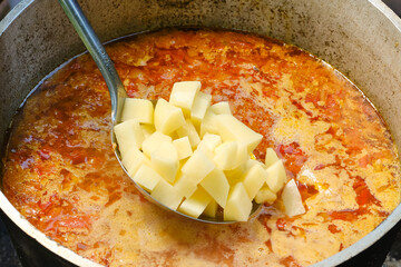 Cooking fish soup or uha in a cast iron cauldron. Delicious fish broth with vegetables. Chef puts chopped potatoes into the soup with a spoon. Shallow depth of field. 