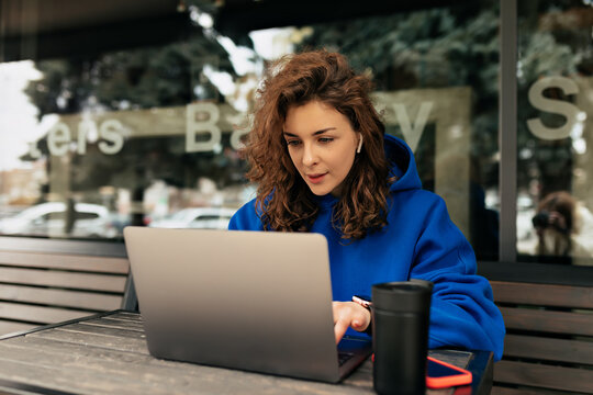 European pretty woman with curly hairstyle wearing blue shirt working on laptop outdoor. Stylish adorable girl sitting on summer terrace with laptop and coffee