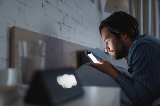 Side View Of Young Man Using Mobile Phone With Blank Screen On Bed At Night