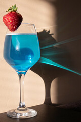 Glass with blue champagne and strawberries on top. Blue sun glare from a glass on a beige wall. vertical photo