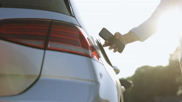 Unrecognizable male unplugging electric car from charging station. Man is unplugging in power cord to an electric car at sunset. Man charging electric car at charging station using smart phone app