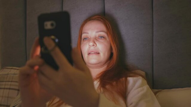 a red-haired emotional woman suffers from insomnia and lies in bed with a phone and looks at websites on the Internet