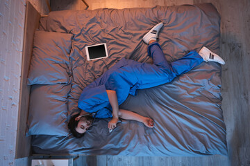 Top view of sleepwalker in doctor uniform sleeping and holding stethoscope near digital tablet on bed at home