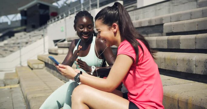 Two sporty women listening to music through earphones and sitting on stairs outside. Young athlete friends taking a break from jogging enjoying, songs on phone. Trendy sportswomen dancing and singing