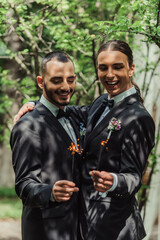 Fototapeta premium cheerful gay newlyweds in formal wear with boutonnieres holding sparklers in green park.