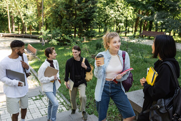 Positive interracial students with devices and notebooks talking in park.