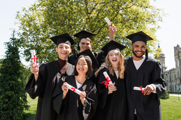 Smiling multicultural bachelors holding diplomas and looking at camera in park.