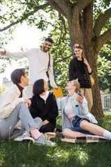 Positive interracial students with backpacks looking at friends on blanket in park.