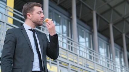 Attractive smiling young businessman in suit eating fast food outdoors, lunch break at office worker