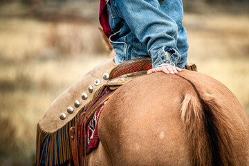 Cowgirl leans back and rests on the hip of her dun horse