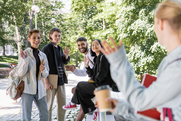 Smiling multiethnic students waving hands at blurred friend in park.