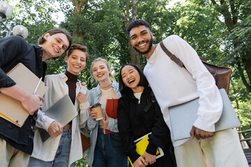 Low angle view of cheerful multicultural students with notebooks and laptops looking at camera in park.