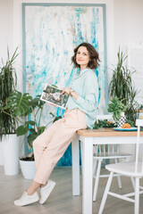 A young woman dressed in pastel colors leafs through a magazine while leaning on a wooden table in...