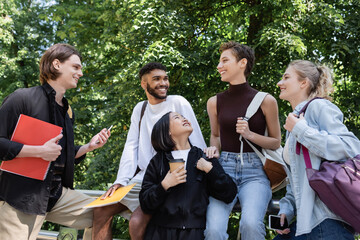Smiling multicultural students with notebooks and smartphones talking in park.