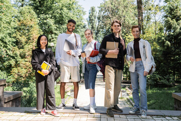 Fototapeta premium Smiling multicultural students with notebooks and laptops looking at camera in park.