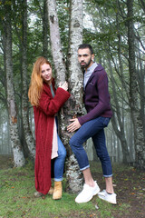 Young woman and man embracing a tree standing between them in a misty forest in autumn. They are wearing colorful warm knitwear and jeans and looking in the camera. Environment protection concept.