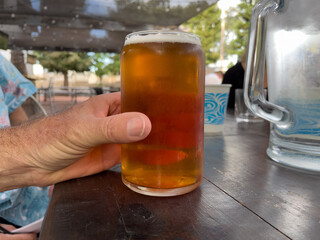 Man's hand grabbing ahold of beer in frosted glass on a table. Ice cold beer on table. Pitcher of ice water sitting on table next to beer. Beer is a medium,golden IPA from brewery
