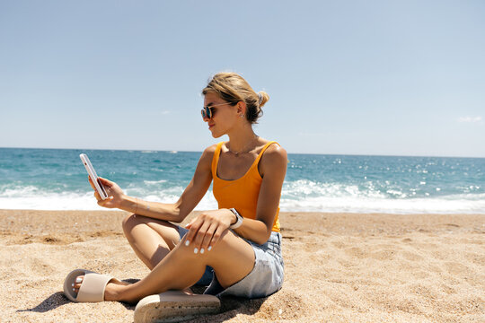 Full-lenght profile photo of pretty slim tanned woman wearing sunglasses and orange t-shirt and denim shorts is holding smartphone while sitting on the beach in sunny hot day 