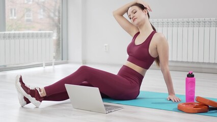 a woman in a burgundy tracksuit with laptop on a mat in a gym. online fitness