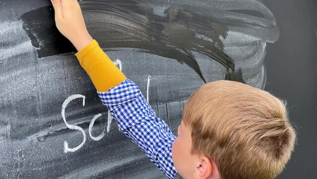 A beautiful Caucasian boy in a plaid shirt erase the inscription Back to school from the blackboard.